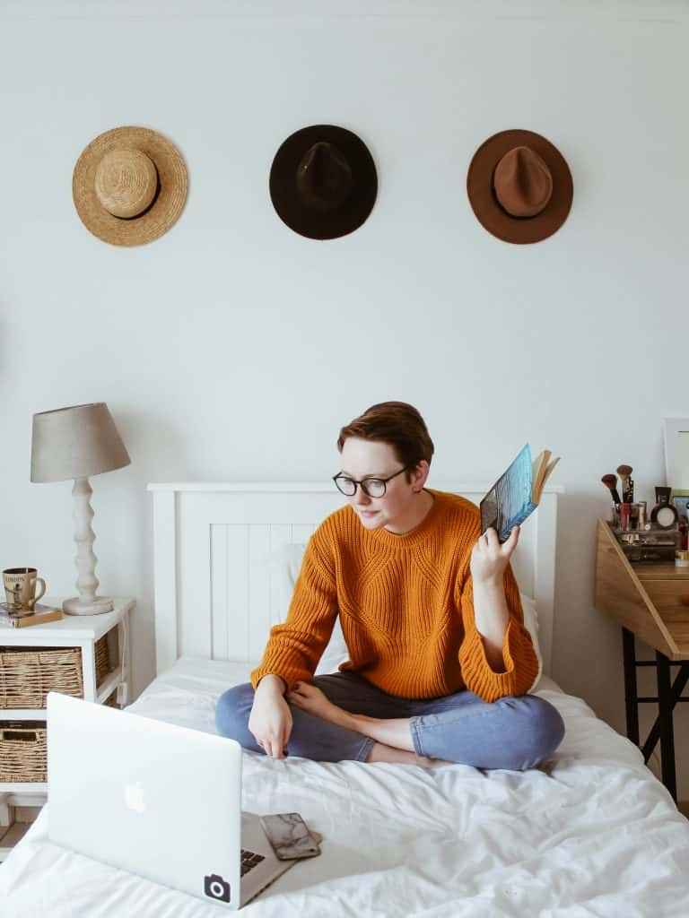 Young woman with glasses reading on bed in a cozy room with hats on wall.